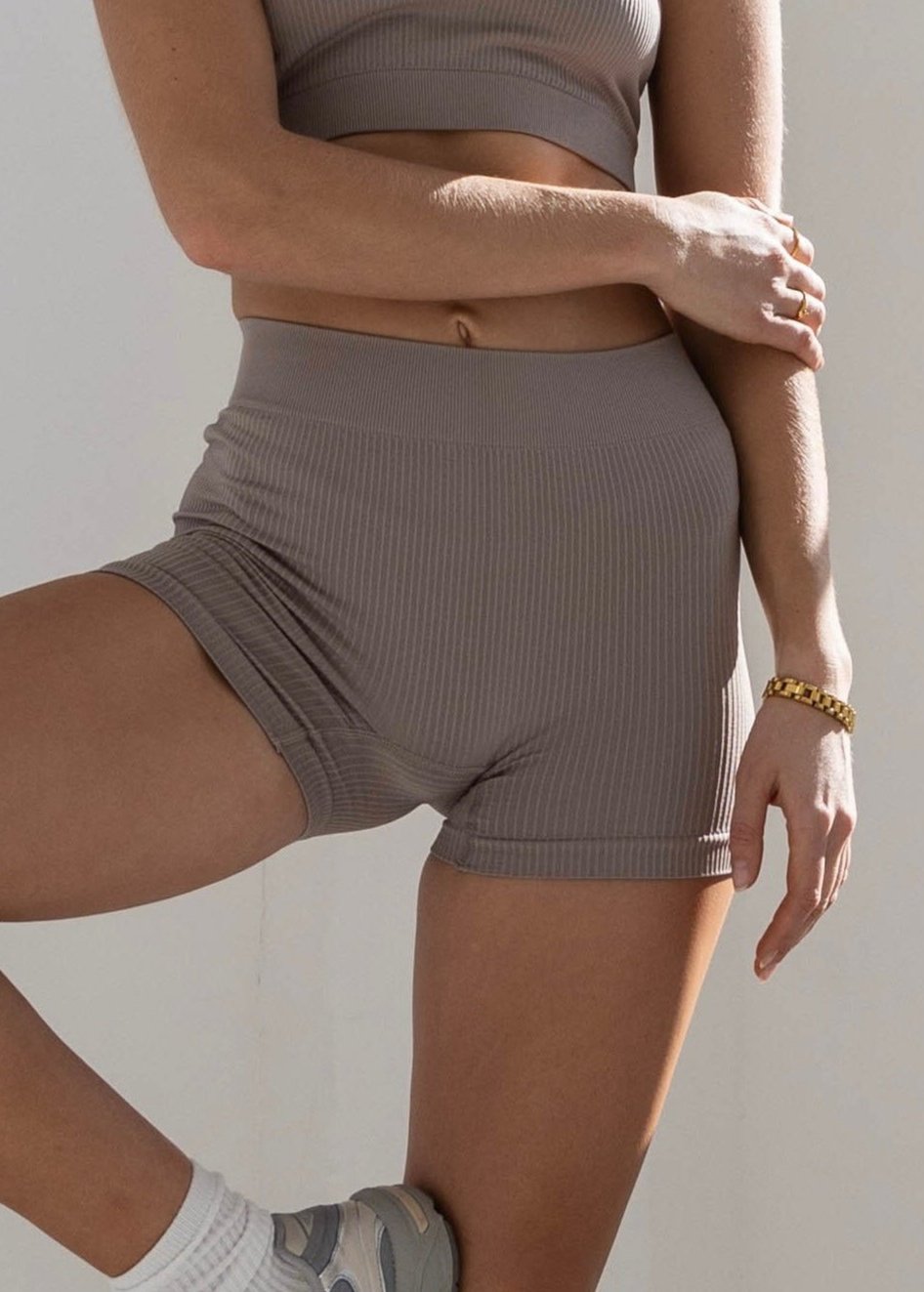 The image feature a close up look of a woman wearing blush form boy-short in ribbed fabric that is sports bra and high waisted shors. she stand with one leg bent and her foot resting against the opposite leg.