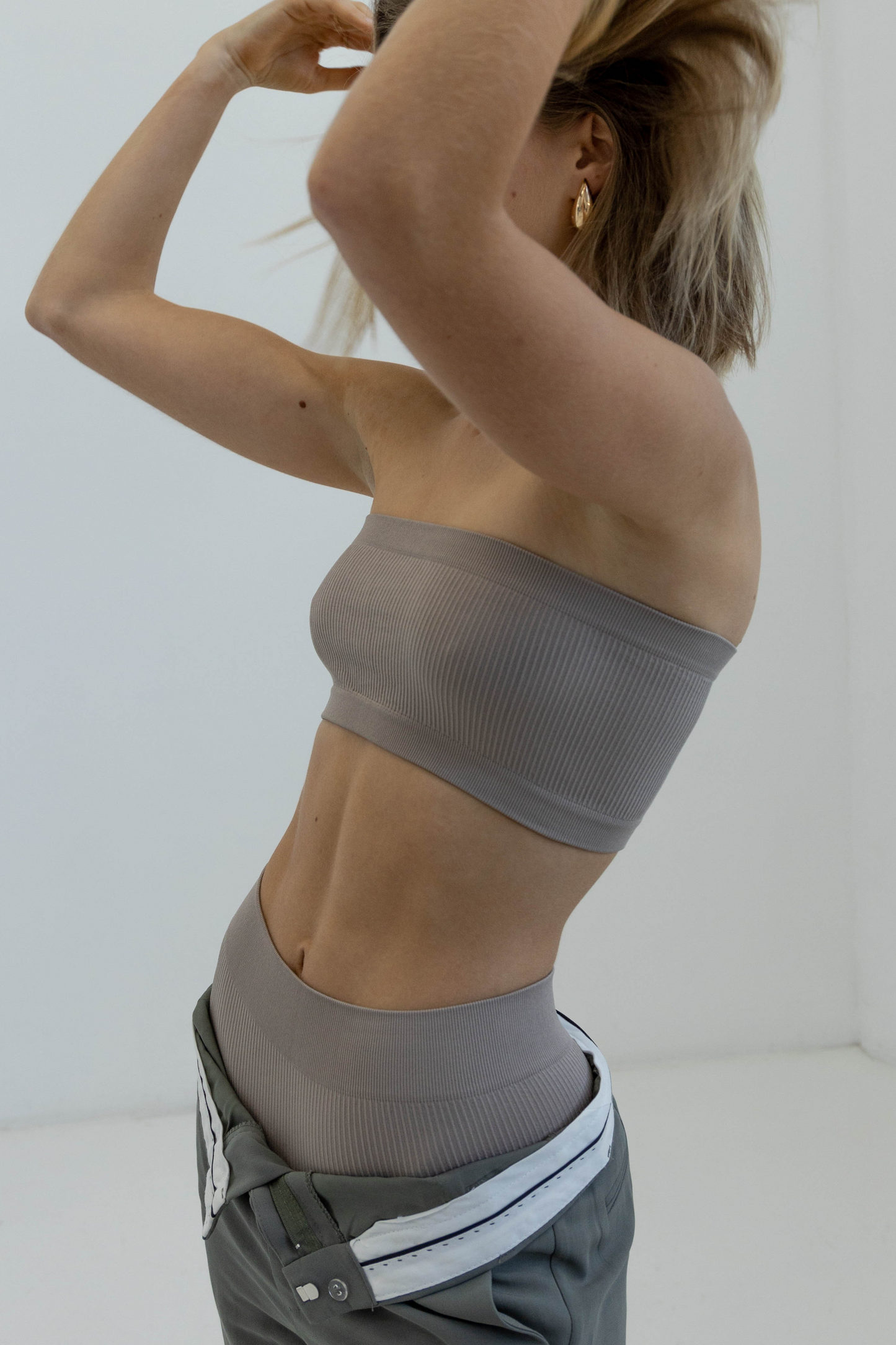 The image display a woman posing with both of her hand on her hair show casing Form blush bandeau facing the side studio, and layer the activewear set with grey trousers to style. that gives