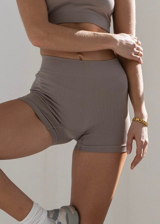 The image feature a close up look of a woman wearing blush form boy-short in ribbed fabric that is sports bra and high waisted shors. she stand with one leg bent and her foot resting against the opposite leg. 
