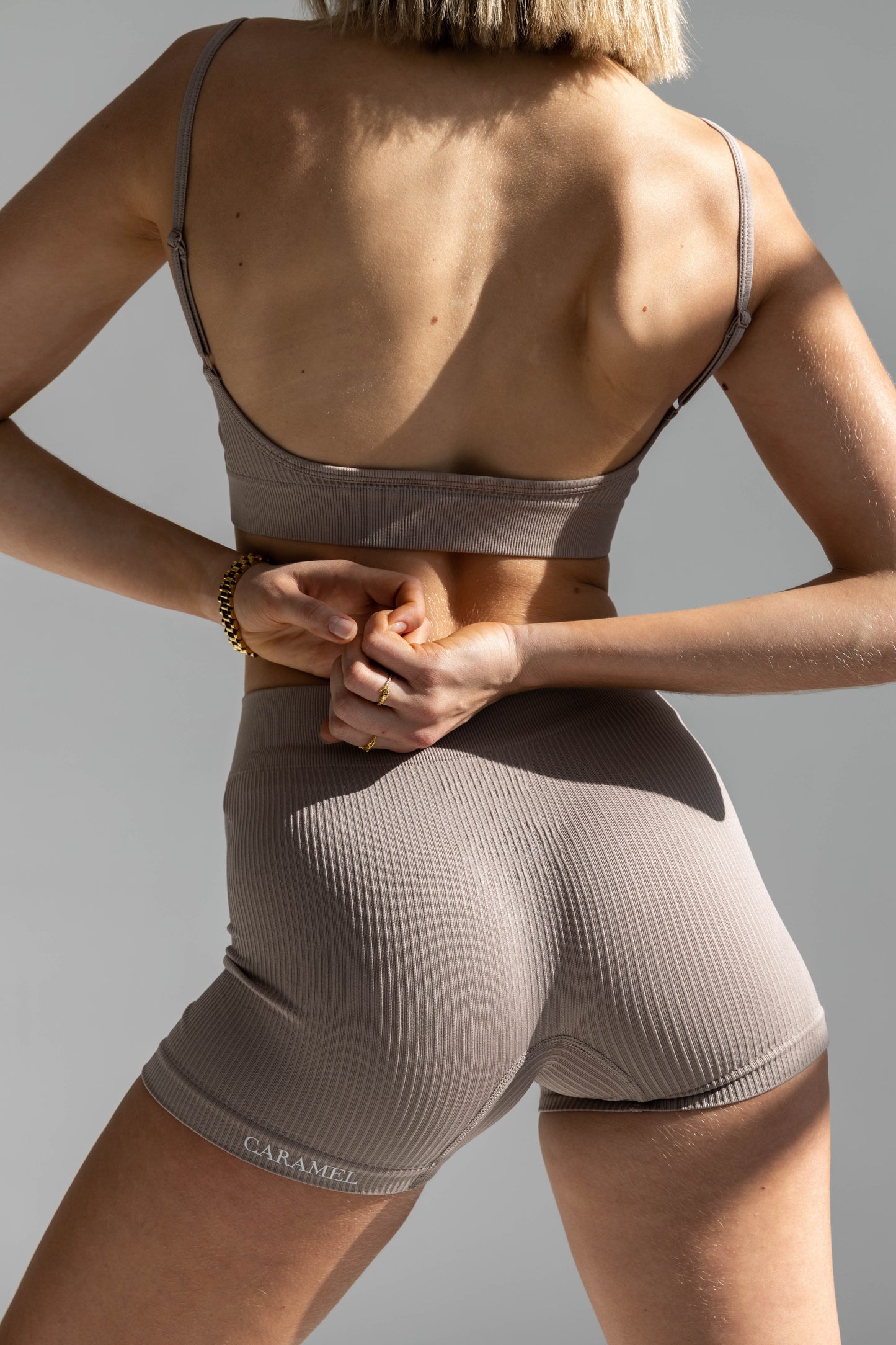 The image shows a woman facing backwards wearing Form blush boy-short set in ribbed material and blush, taupe colour with her one leg bend to one side and her both hands intertwining in the back 