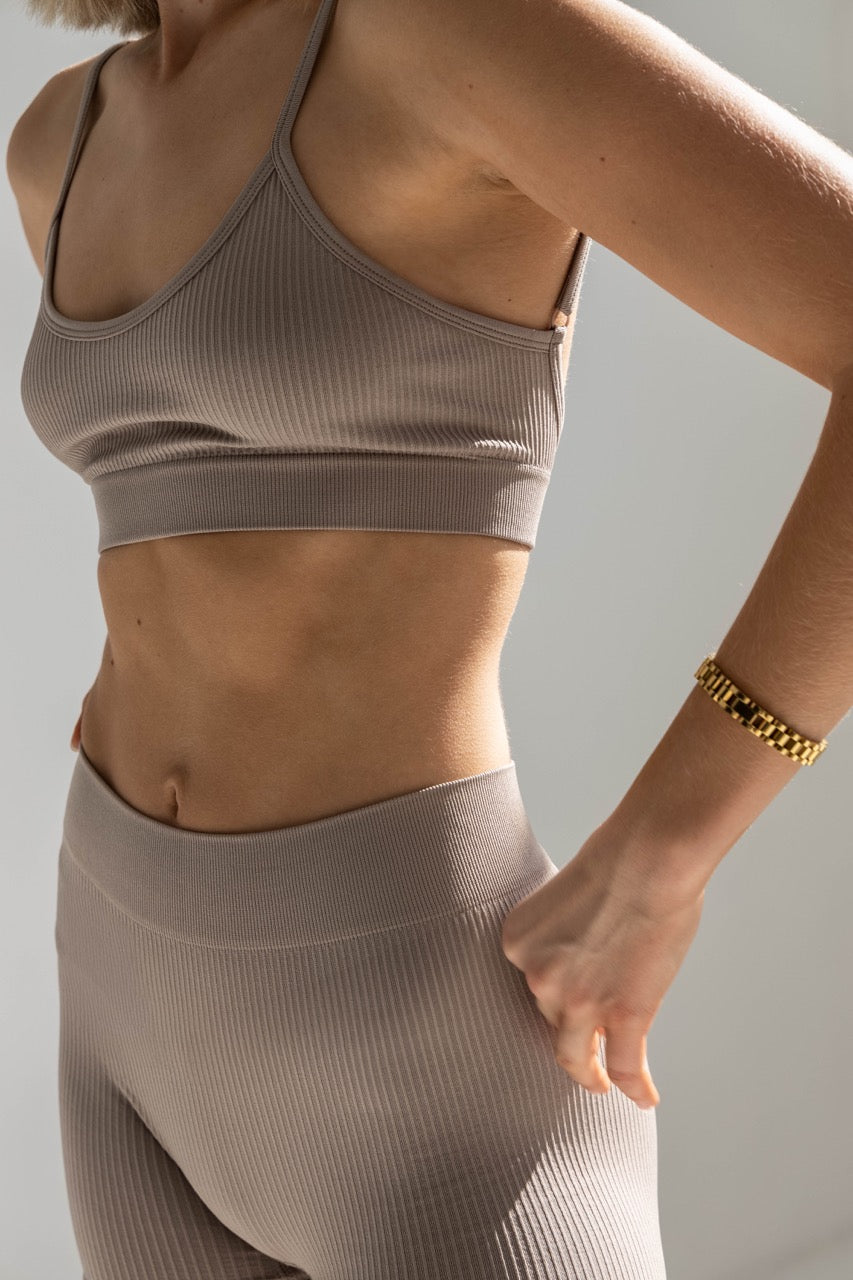 The image shows a close-up of a person wearing a ribbed form bralette and matching high-waisted leggings. The sports bra has a scoop neckline and thin, adjustable straps, with a supportive band under the bust. The fabric appears soft and stretchy, emphasizing comfort and light support. The leggings are high-waisted with a wide waistband, offering a snug yet flexible fit. The person’s hand is resting on their hip, and they are wearing a gold bracelet. 